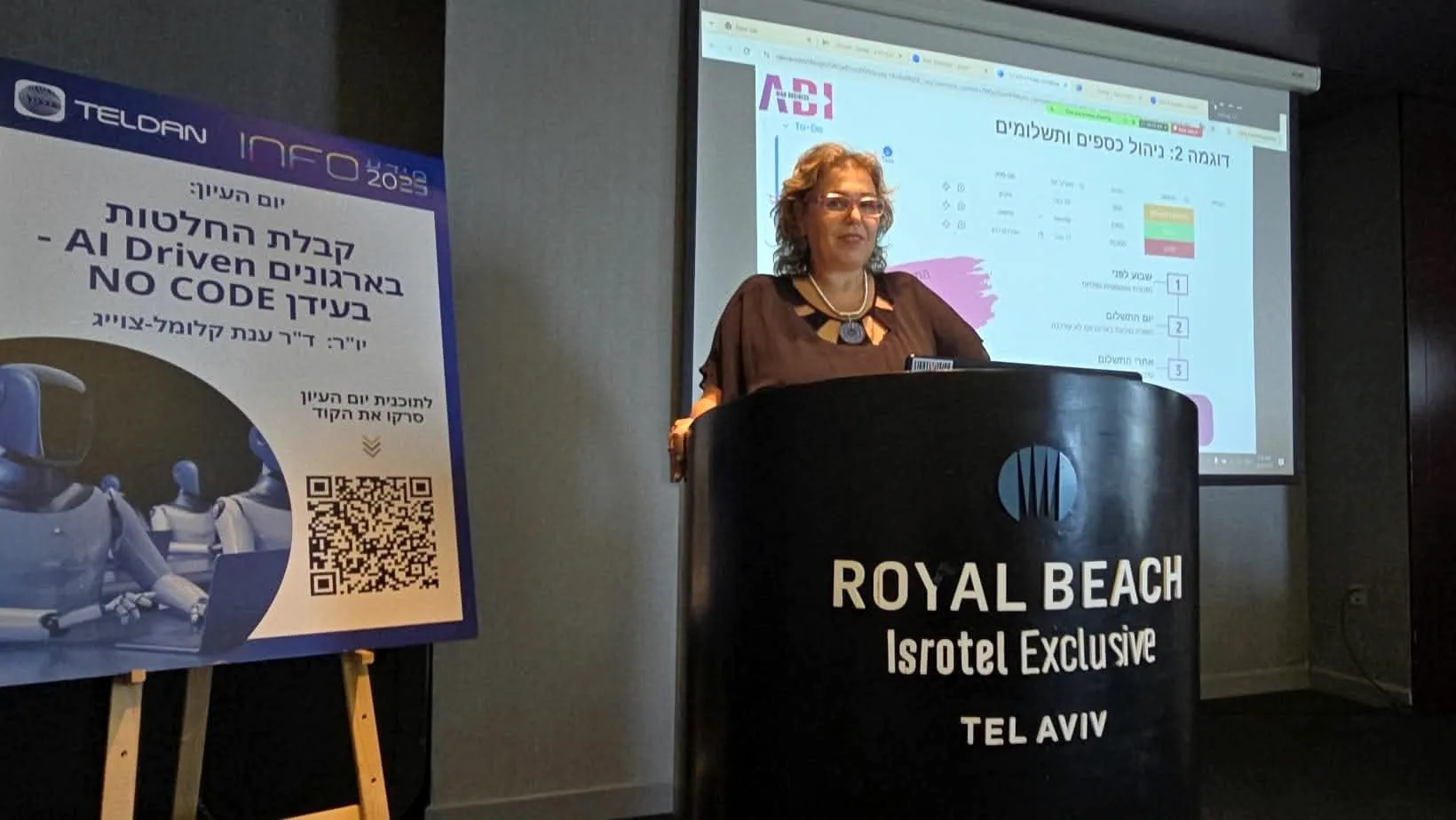 ד״ר ענת קלומל-פרוכצוייג מרצה בדוכן לצד כרזה על קבלת החלטות, בינה מלאכותית ו-No Code