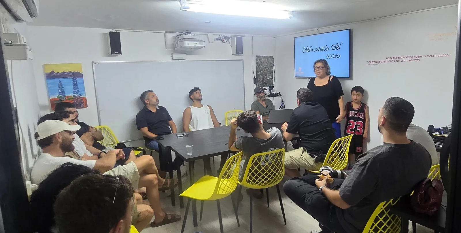 ד״ר ענת קלומל-פרוכצוייג עומדת מול משתתפים סביב שולחן בכיתה עם כיסאות צהובים