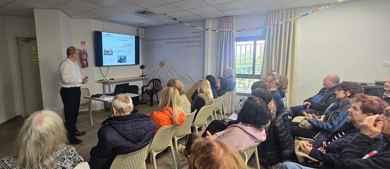 מרצה עומד לצד מסך ומציג מצגת לקהל היושב בכיתה